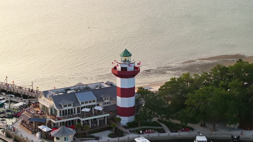 Lighthouse reflecting in harbor water during vibrant sunset over pier, telephoto aerial orbit, Harbour Town Pier South Carolina USA