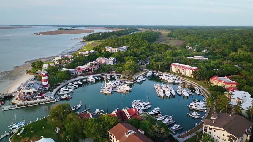 Aerial glide pullback over circular yacht marina passing lighthouse at tip of curved pier surrounded by calm harbor, Harbour Town Pier South Carolina USA