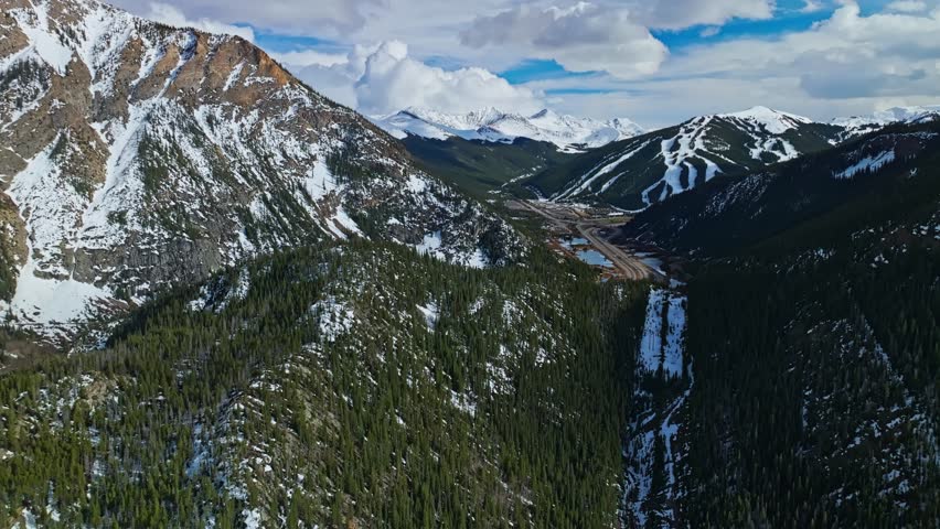 Flying over rocky valley in Frisco Gulch with thick pine trees and morning sun, aerial pullback with highway in distance, snow spring on mountains