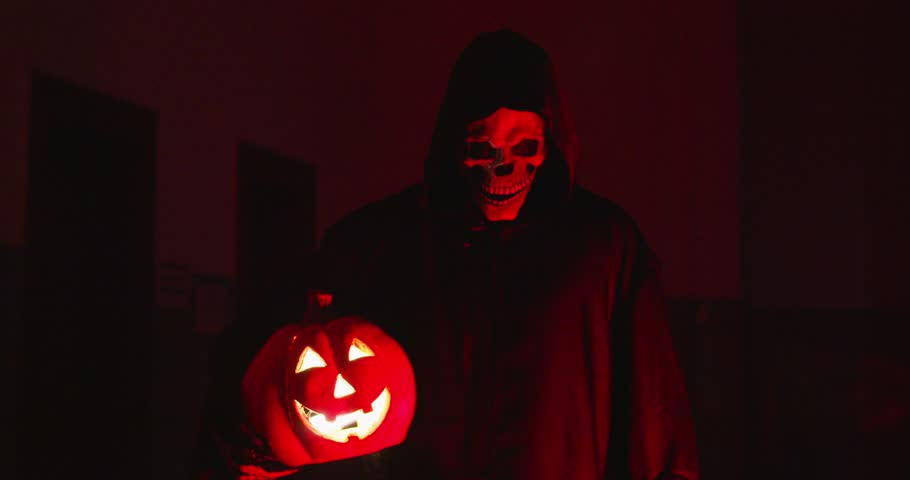 Dark macabre figure in scary Halloween costume. Sinister Death Reaper, nightmare spirit, or eerie monster disguised in cloak holds glowing Jack o lantern and shows shush shhh shut up silence gesture