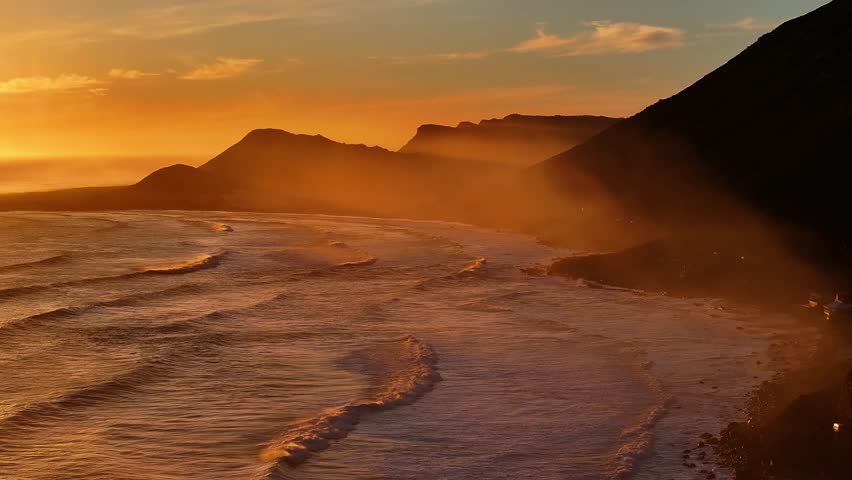 Aerial view of serene sunset over waves and coastal mountains with clouds, Scarborough, Western Cape, South Africa.
