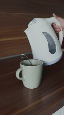 Unrecognizable girl pouring hot water from electric kettle into mug on kitchen counter