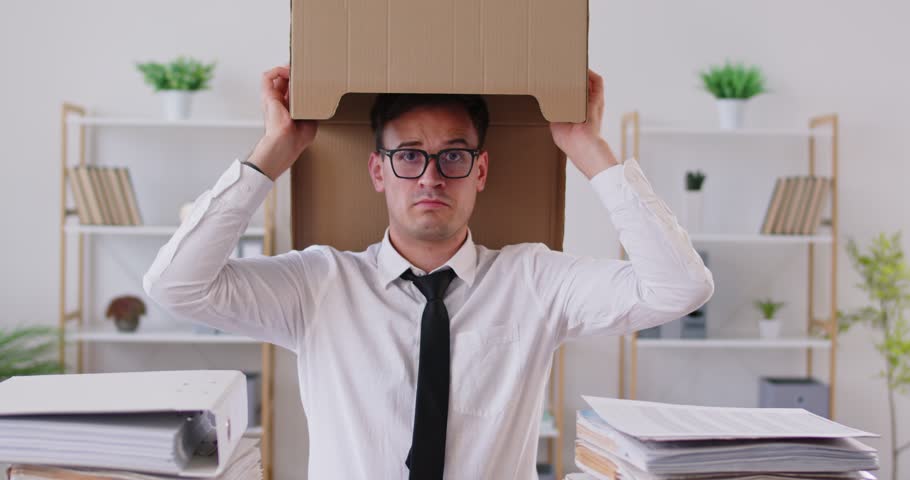 Sad company employee in eyeglasses sitting at desk, hiding his head in cardboard box, trying to escape from stress. Funny frustrated male worker taking break from work, fooling around and dancing.