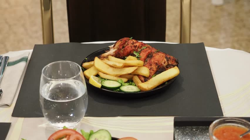 Tandoori chicken legs are served on a black plate with chips, cucumber and lemon slices, accompanied by a glass of water and a small bowl with tomato and cucumber slices and spicy sauce