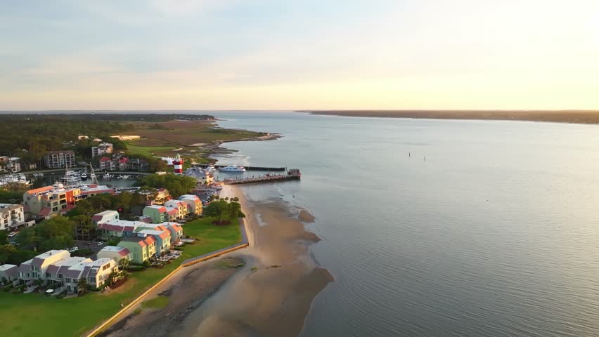 Glide over boats and lighthouse at harbor town pier as sun dips near horizon, panoramic aerial of Harbour Town Pier South Carolina USA