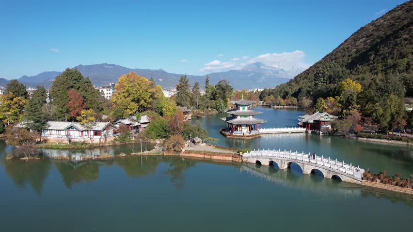 A serene view of Black Dragon Pool in Lijiang, with calm waters reflecting ancient architecture. Snow-capped mountains rise in the distance. Trees sway gently in the breeze. A peaceful moment in natur