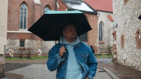 A man in a denim jacket and hoodie walks through a cobblestone street on a rainy day, holding a dark green umbrella. The scene is set in a charming European old town with historic stone buildings. - Powered by Shutterstock - Get 15% off with code: PIKWIZARD15