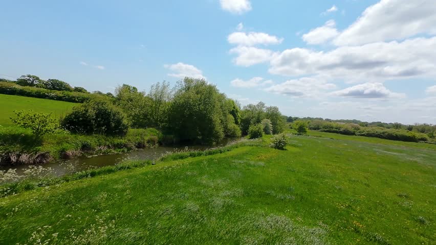 A drone view of a small stream winding through lush green fields, weaving a natural path across vibrant summer countryside
