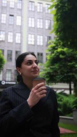 Stylish confident lady in black office suit seated in business center yard, drinking takeaway coffee with thoughtful look. Concept of career woman, morning routine, urban confidence.