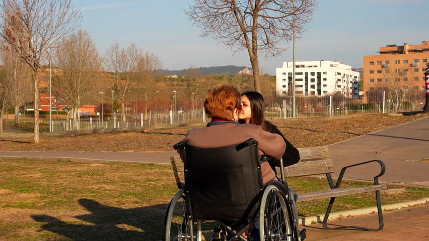 Rear shot of an elderly woman in wheelchair talking with granddaughter on park bench on sunny day