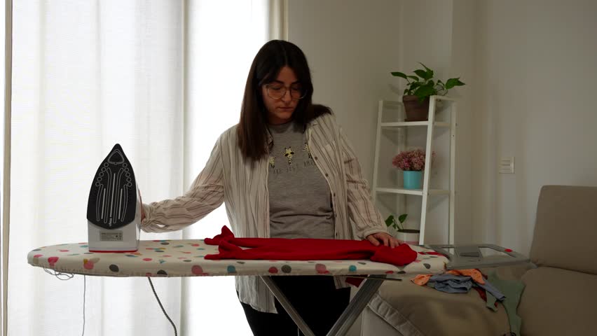 Mother folds red Christmas baby pajamas after ironing, preparing for newborn