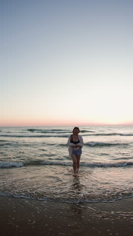 Vertical shot. Young plus size woman enjoying a walk on the beach during sunset. Slow motion of female feet walking barefoot on the beach at golden sunset leaving footprints in the sand. 