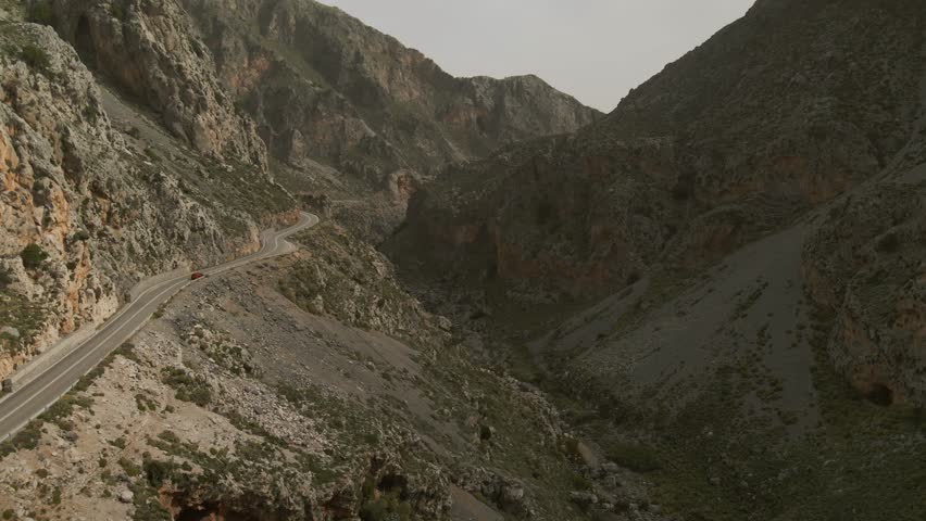 Aerial view of a narrow paved road snaking along steep cliffs and rocky landscape in Kourtaliotiko Gorge, Crete, Greece, under a hazy sky with minimal vegetation