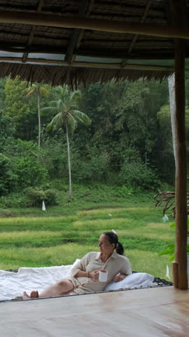 Vertical video. Sitting in a hammock, a woman drinks her coffee while gazing at lush greenery and rice terraces, slow motion