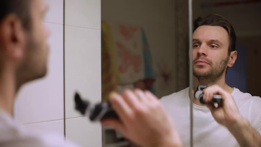 A man in a white T-shirt trims his beard with an electric shaver while looking into a bathroom mirror, focusing on grooming and personal hygiene during a morning routine.
