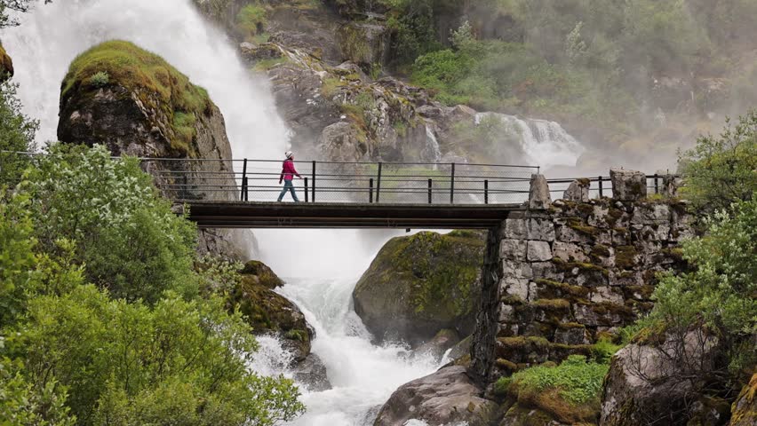 Woman hiker crosses a bridge over the Kleivafossen waterfall on the way to Brikdalsbreen Glacier in Norway.
