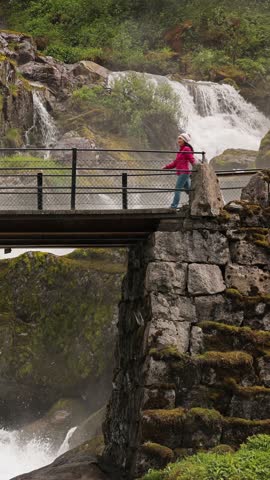 Woman hiker crosses a bridge over the Kleivafossen waterfall on the way to Brikdalsbreen Glacier in Norway. Vertical Video.
