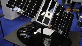 CubeSat model with glossy black solar panels mounted on rotating base displayed at science expo demonstrates compact satellite technology for space research communications and orbital innovation - Powered by Shutterstock - Get 15% off with code: PIKWIZARD15