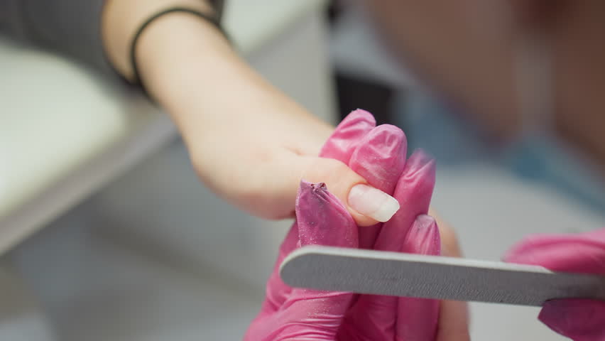 Nail technician wearing pink gloves carefully files client fingernail using white OPI nail file, focusing on shaping nail tip during manicure process in clean, professional salon environment