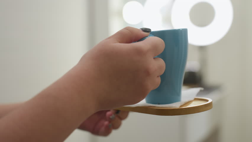 Close-up of beautician holding blue tea cup with care and gently placing it on table in softly lit salon, creating warm and attentive service atmosphere for relaxing beauty treatment experience