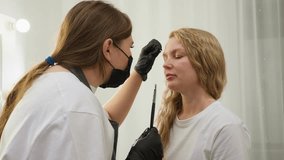 Beautician wearing black gloves and mask applies cream to client eyebrow with brush while gently shifting client s hair aside. Client with blonde wavy hair sits calmly in bright, clean beauty salon - Powered by Shutterstock - Get 15% off with code: PIKWIZARD15