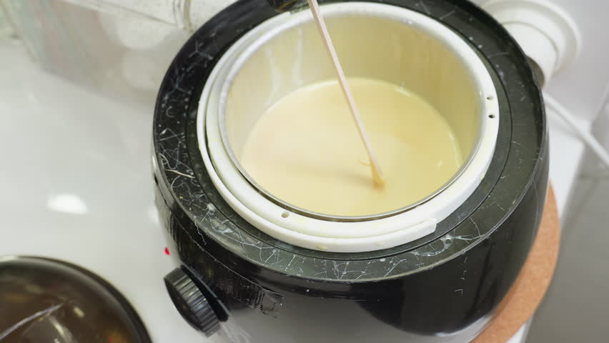 Close up of beautician wearing black glove using wooden stick to stir creamy substance inside electric heater, likely preparing for cosmetic treatment or waxing in professional salon setup