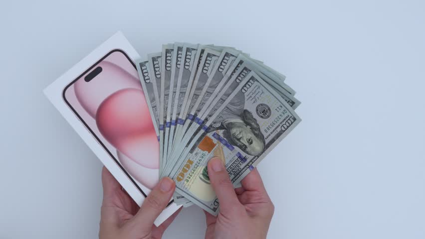 Ottawa, ON Canada - 06.04.25: A pair of hands holding new smartphone iPhone box and stack of hundred-dollar bills. Buying new phone, wealth and technology.