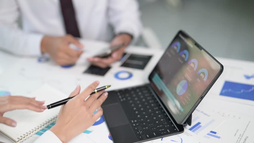 Two people are working on a laptop and a notebook. One person is writing on the notebook while the other person is looking at the laptop. The laptop has a screen displaying graphs and charts