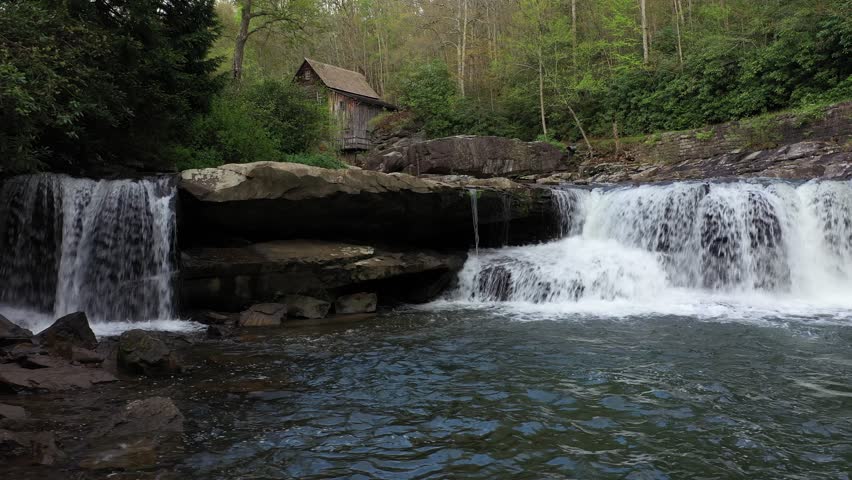 Experience the beauty of spring in West Virginia, showcasing the iconic grist mill surrounded by lush greenery and a serene waterfall cascading into a creek.