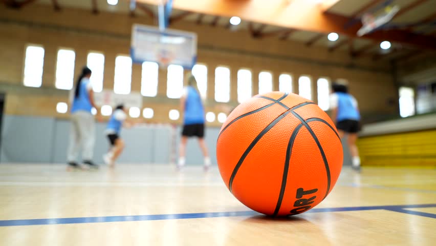 Orange basketball resting on hardwood court with blurred players practicing in background during dynamic training session