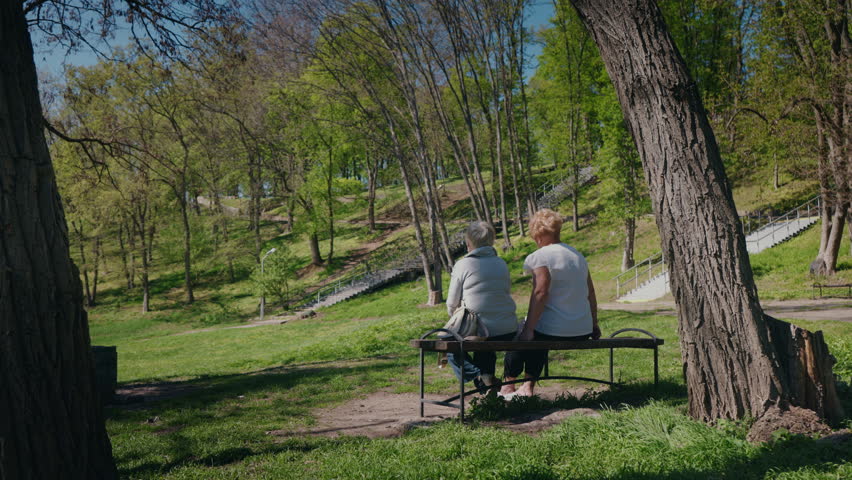 Two elderly women sit on a bench in a sunny park, relaxing and chatting while enjoying the peaceful spring scenery around them. High quality 4k footage