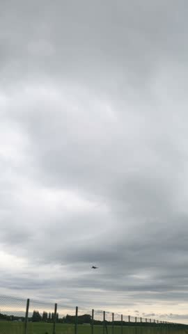 Vertical screen low-flying military jet approaches over fence under heavy gray sky capturing the raw tension of wartime readiness and silent anticipation on the ground