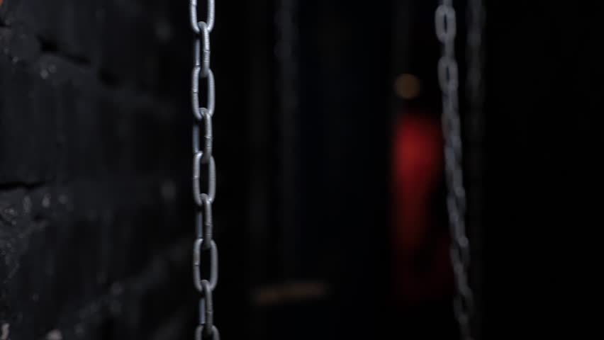 Close-up of hanging chains in a dark room with multicolored light bulbs in the background. The chains swing from side to side. 