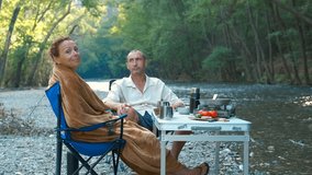 Couple enjoying breakfast by river during camping trip. Happy couple relaxing and having breakfast by the river, wrapped in a blanket on camping chairs next to a portable table with food and drinks - Powered by Shutterstock - Get 15% off with code: PIKWIZARD15