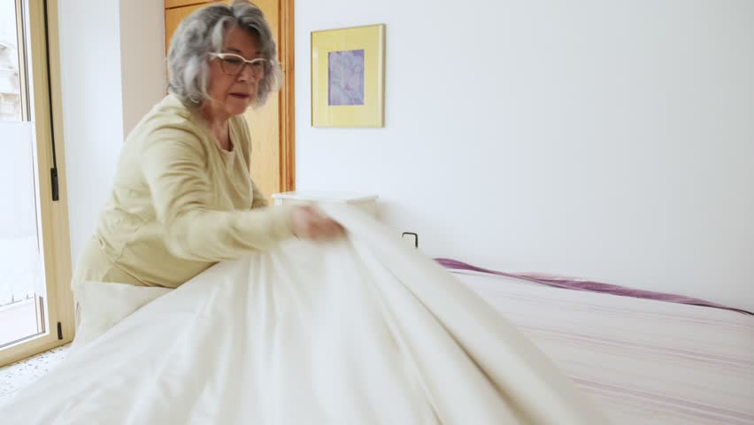 An elderly woman tidies up her bedroom by making the bed, adding a personal touch to the space. This short segment highlights the simplicity and beauty of daily routines in a serene environment.