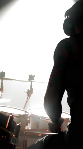 Cinematic shot on stage of concert hall, talented drummer playing drum set, silhouette against bright light of soffit. Brawny male arms and torso, man playing music and beating rhythm, vertical video