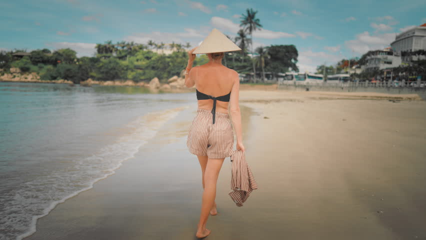 Graceful woman in bikini, shorts and conical hat walking on seashore in Vietnam. Back view of lady strolling alone on beach, happy and carefree female vacationer enjoying simple life, traveling