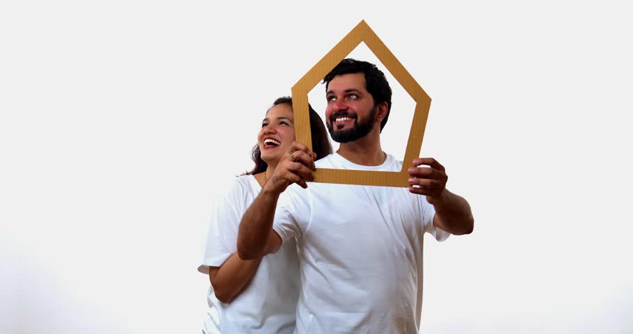 Couple looking through home cutout, standing isolated against white background, Indian Asian young man and woman smiling at camera while holding house-shaped stencil, promoting real estate investment