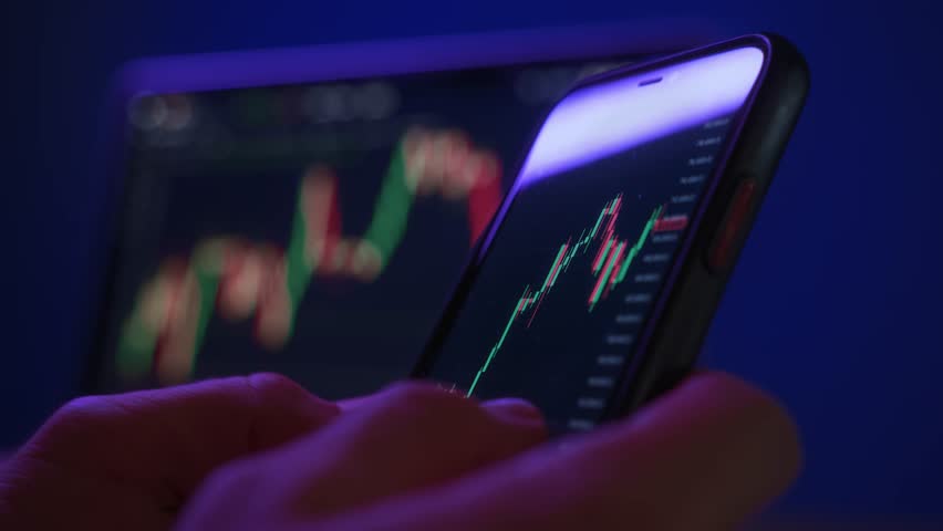 Close up, trader or investor using smartphone for real-time stock market monitoring with candlestick chart in a modern tech setup with neon light. Trading app with crypto graphs on screen