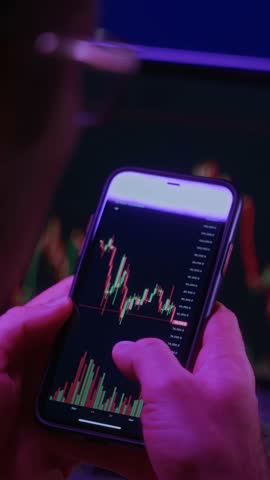 Over the shoulder view of a trader closely examining cryptocurrency charts on a smartphone via a trading app, with stock market data displayed on a laptop in the background. Vertical video