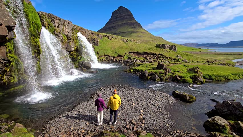 A couple stands hand in hand, admiring the stunning Kirkjufell mountain in Iceland. The dramatic waterfalls flow nearby, surrounded by lush greenery under a clear blue sky. Perfect for nature lovers.