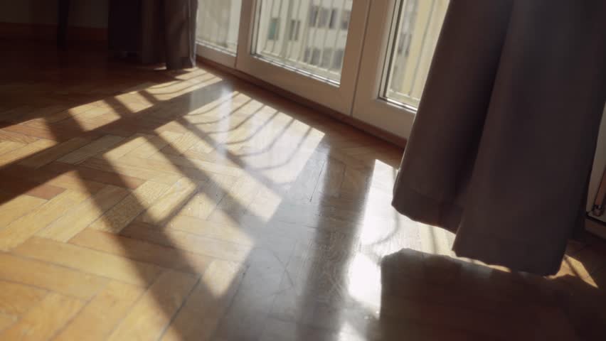 Shadows of sunlight entering from balcony window in apartment during daytime. Beautiful sun shining through as camera pulls away. Bedroom or hotel in early morning. Parquet floor inside European room