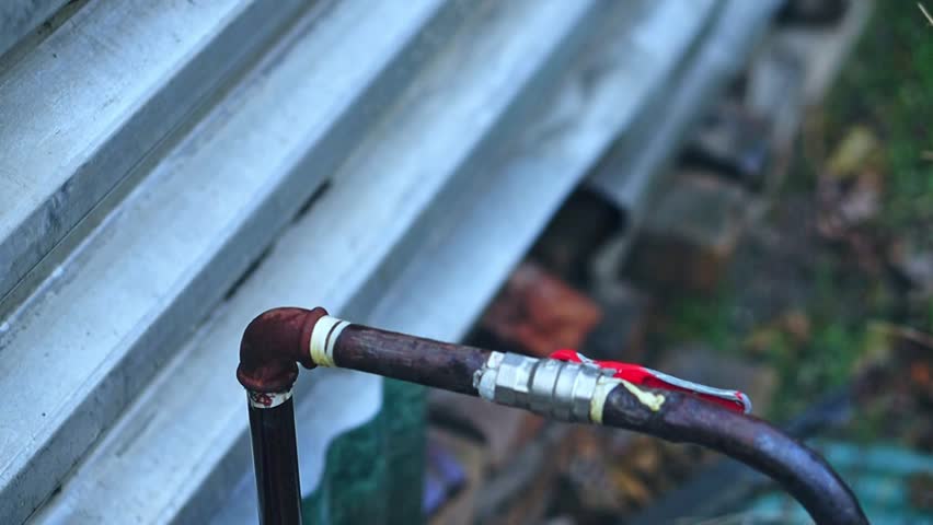 A white man's hand closes a red-handled ball valve and a stream of water starts to burst from the corner, signaling a leak and a rupture in the rusty pipeline. Plumbing equipment failure, broken