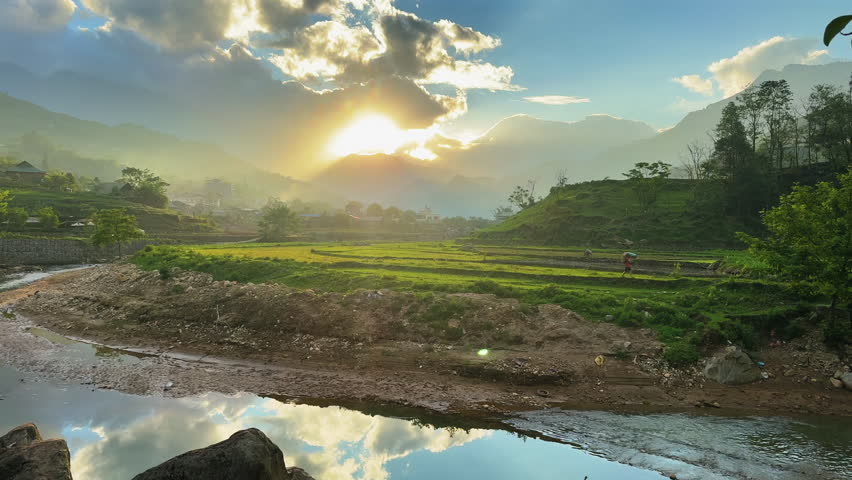 Static wide shot of terraced fields, mountain silhouettes, and river near Tả Van, Sa Pa, as sun sets behind clouds, reflected in water, peaceful rural scenery, no people.