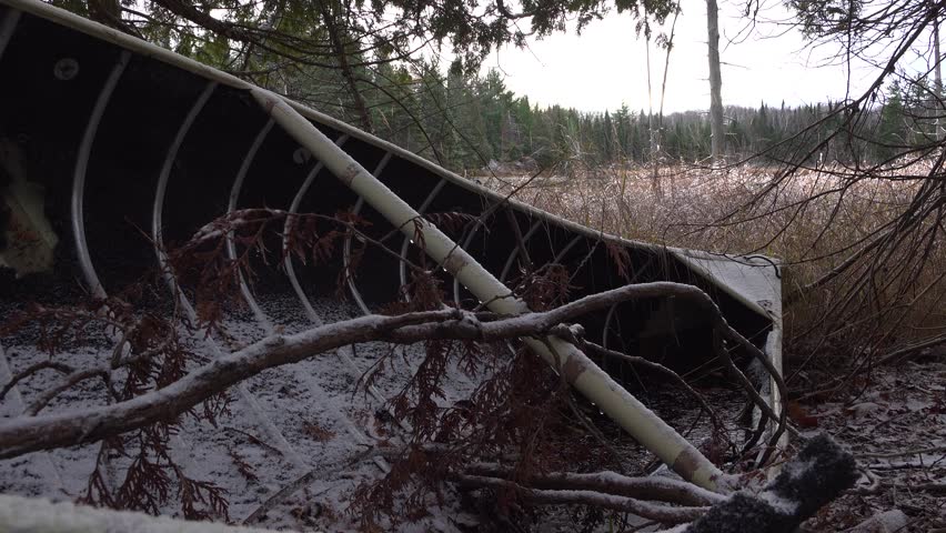 An abandoned canoe rests amidst a snowy shoreline with trees, winter foliage and a forest setting providing an atmosphere of quiet and solitude.