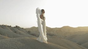 Spiritual woman in white dress and angel wings standing in desert landscape, raising hand against golden sunset light, embodying divine freedom and ethereal beauty - Powered by Shutterstock - Get 15% off with code: PIKWIZARD15