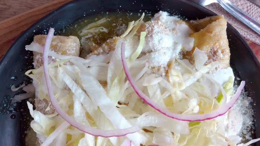 Overhead shot zooming out from a plate of Mexican enchiladas verdes, topped with lettuce, onion, and cheese.