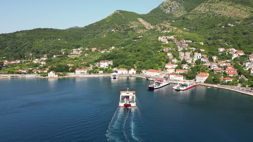 Aerial view of a passenger ferry sailing in the Bay of Kotor against the backdrop of a picturesque mountain coastline and a seaside town on a sunny day