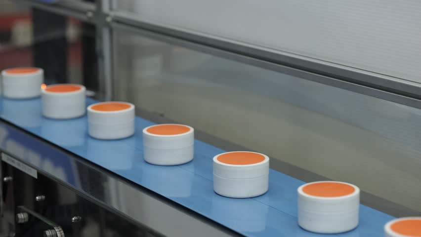 Plastic jars with orange lids moving on a blue conveyor belt inside a sterile manufacturing plant, highlighting precision and automated packaging process in production.