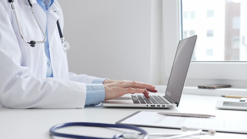 Doctor woman is typing on a laptop with a stethoscope, medical record, and pen on the desk in a bright medical office, showcasing healthcare technology and medical documentation. Medicine concept
